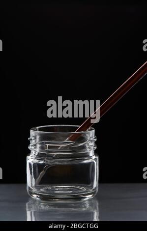 Brush with the maroon handle inside an empty transparent glass jar on a black background. Stock Photo