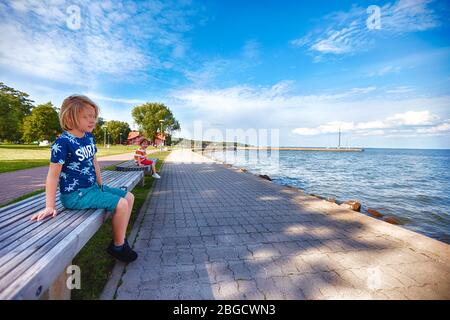 cute kids are sitting on benches at the seaside embankment. Joudkrante, Lithuania Stock Photo