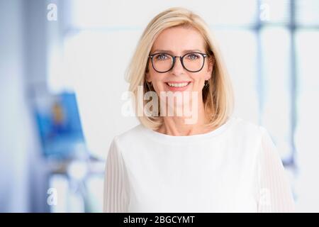 Close-up portrait of beautiful blond businesswoman standing in the office while looking at camera and smiling. Stock Photo