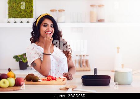 beautiful young woman preparing meal, biting a vegetable during cooking Stock Photo