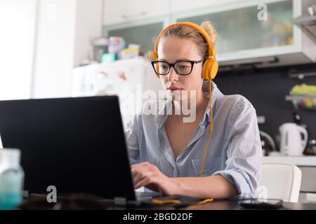 Female freelancer in her casual home clothing working remotly from her dining table in the morning. Home kitchen in the background. Stock Photo