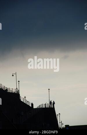 Silhouette of promenade at Cromer showing stark outlines of two men in conversation with street lights and fence against a brooding sky Stock Photo