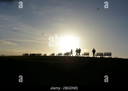 The sinking sun creates a striking silhouette of four figures walking past a row of bench seats on a  hill with halo like sun effect and pale blue sky Stock Photo