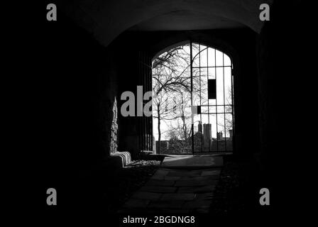 Monochrome image showing a dark passage way leading to a large metal gate set in the wall with one side open and sun coming in Stock Photo