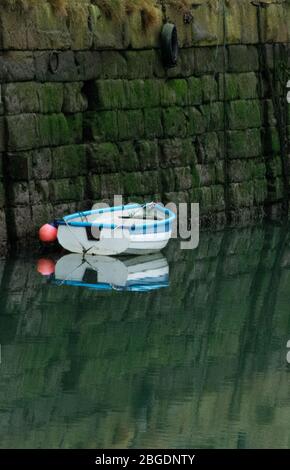 Small blue and white rowing boat with orange buoy tied up against harbour wall with still water and a clear mirror reflection Stock Photo