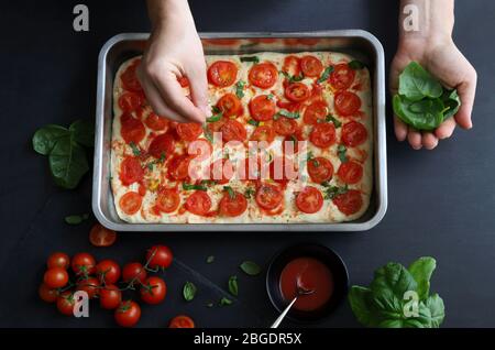 Cooking concept. Homemade italian food. Woman hand decorating pizza with fresh basil leaves on dark background. Top view. Stock Photo
