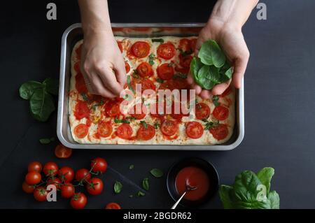 Cooking concept. Homemade italian food. Woman hand decorating pizza with fresh basil leaves on dark background. Top view. Stock Photo