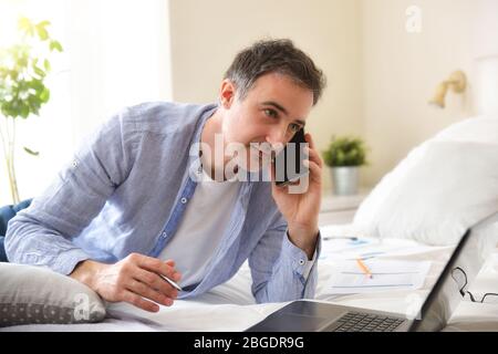 Businessman dressed casually in shirt and jeans working with a laptop lying on a bed with documents Stock Photo
