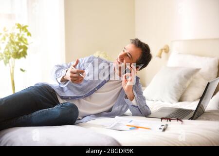 Business man in shirt and jeans talking on the phone leaning with his elbow resting on a bed with documents and laptop Stock Photo