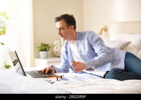 Concentrated businessman dressed casually in shirt and jeans working with a laptop lying on a bed with documents Stock Photo
