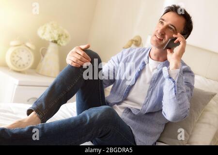 Businessman dressed casually in shirt and jeans talking on the phone sitting along a bed detail Stock Photo