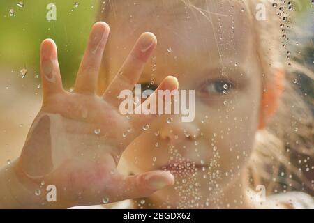 little girl looks out the wet window. Raindrops on the glass. Forest outside the window. Stock Photo