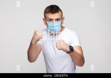 Portrait of serious young man in white shirt with surgical medical mask standing with boxing fists, looking at camera and ready to attack or defence. Stock Photo