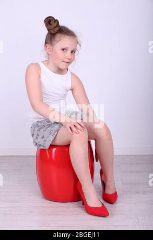 Beautiful small girl in big shoes sitting on leather ottoman on wall background Stock Photo