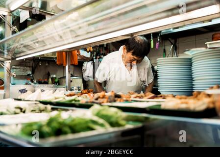 Singapore, Oct 2019: Old man in apron preparing and cooking traditional chinese food at street food stall at a hawker centre. Asian cuisine Stock Photo