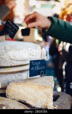 market stall with italian hard cheese sliced and open, alp cheese label (toma alpeggio) on foreground and people who buy the cheese on background Stock Photo
