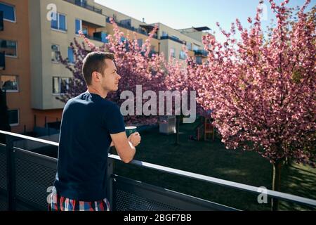 Spring morning at home. Young man with cup tea or coffee on balcony. Stock Photo