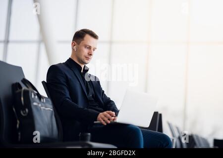 Man with suitcase sitting in airport waiting area while listening music using airpods Stock Photo