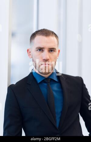 Middle-aged man standing with folded arms leaning against a windowsill turning to look at the camera with a pensive expression Stock Photo