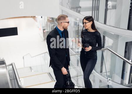 Beautiful business woman and man their holding a tablet in hands and talking to each other while standing at staircase at modern office building. Stock Photo