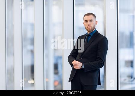 Middle-aged man standing with folded arms leaning against a windowsill turning to look at the camera with a pensive expression Stock Photo