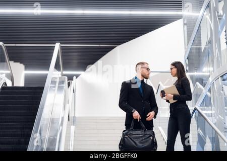 Business couple on a stairs talking and smiling. Man wearing suit and glasses with a bag talking with a woman holding tablet computer. Stock Photo
