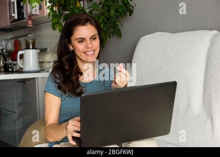 Brunette girl shows a finger gesture of love in Korean and looking at her laptop making video call in her apartment. Fun greeting online. Using Stock Photo