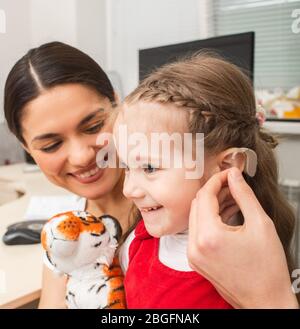 Child is not afraid that she needs to use a hearing aid on her ear. Mother of child is very pleased that her daughter will hear the world. Deafness tr Stock Photo