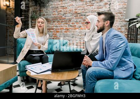 Group of three focused multiethnic male and female business people in formal clothes gathered around laptop on the table in modern office and Stock Photo