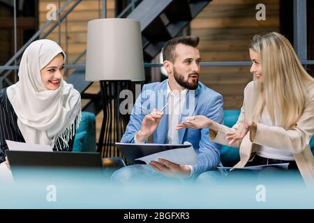 Pretty muslim business lady in hijab, Caucasian man and attractive blond woman, sitting in the modern loft office room, smiling and discussing key Stock Photo