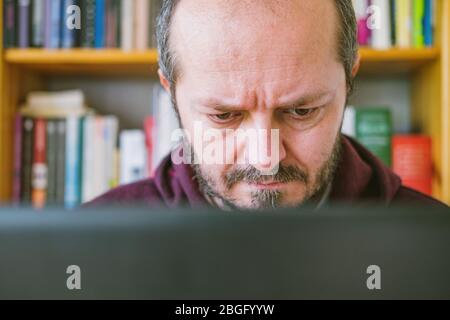 Adult man working from home office. Bearded man dedicated working online from home on computer laptop, book shelves behind him. Close up. Stock Photo