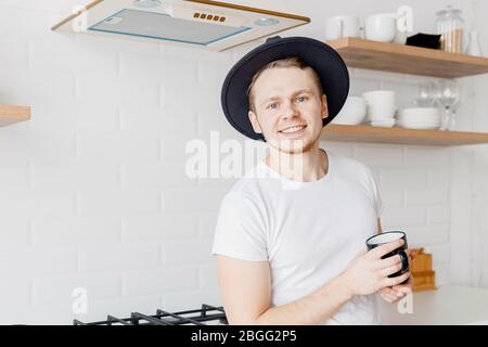 Portrait young man in hat smiles drink fresh coffee in ceramic black mug breakfast. Bright kitchen background Stock Photo