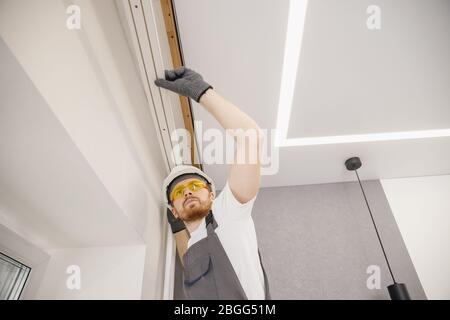Male builder carpenter installs curtain on ceiling, bottom view Stock Photo