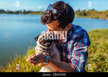 Man hugging and playing with pug dog in park. Man walking with pet by spring lake. Friends enjoying nature Stock Photo