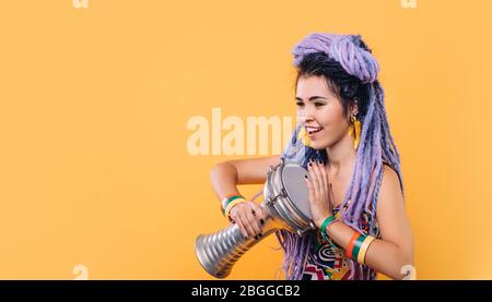 Hipster woman with violet dreadlocks and colored clothes playing on ethical mini drum on yellow background Stock Photo