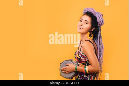 Rastafarian woman with violet dreadlocks and colored clothes playing on ethical mini drum on yellow background Stock Photo