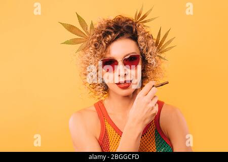 Hipster woman smokes marijuana through a special smoking pipe. A woman contributes to legalization of cannabis. Portrait on yellow background Stock Photo