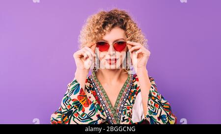 beautiful woman with curly haircut holds her trendy red glasses with her hands on violet background Stock Photo