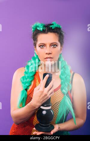 Big glass bong in the hands of a hipster woman. Woman smokes cannabis. Female hairstyle with green kanekalon on a violet background. Legalization of h Stock Photo