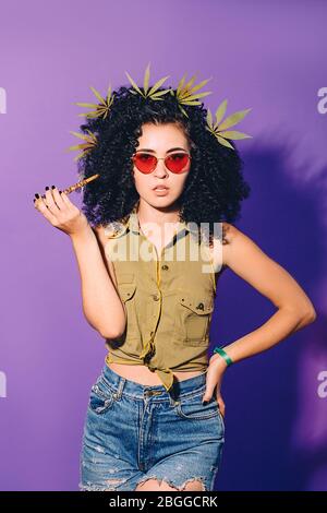 Hipster woman smokes marijuana through a special smoking pipe. A woman contributes to legalization of cannabis. Portrait on Violet Background Stock Photo