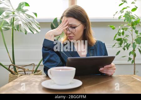 Beautiful mature woman in pajamas reading news, social networks, mail in digital tablet. Morning portrait of middle-aged female at home at table with Stock Photo
