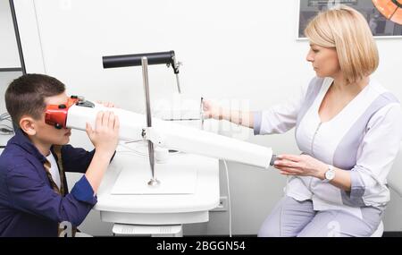 Boy looking into device to stimulate vision. Female ophthalmologist monitoring treatment of visual acuity using pleoptic methods Stock Photo