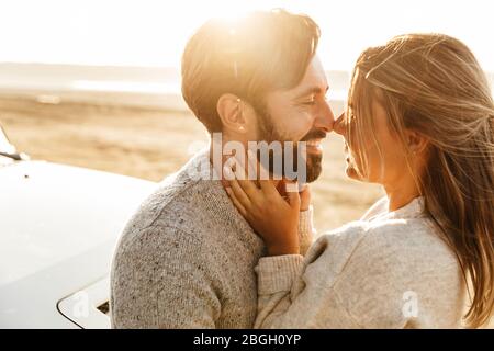 Close up of a happy young couple in love embracing while leaning on a car at the sunny beach