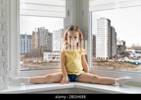 beautiful little girl sitting on the windowsill and smiling, looking at the camera. outside the window is rain and cityscape. stay home with coronavir Stock Photo