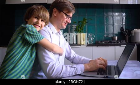 A man works in the kitchen at a laptop with his children at home. Father with son. Remote work at home during the coronavirus quarantine period Covid 19. Stock Photo