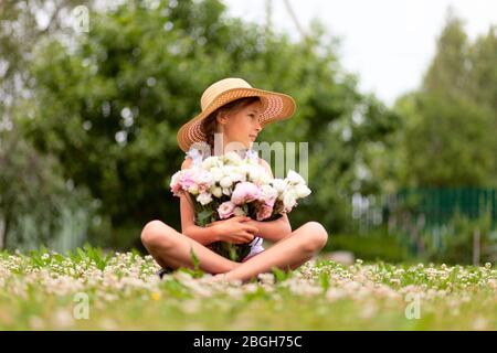 A bouquet of pink and white peonies in the hands of a girl sitting in a flowering meadow. A child in a straw hat with wide brim on a background of green trees. Sunny day. Stock Photo