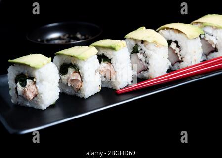 Sushi roll with avocado wrap, grilled salmon, Philadelphia cheese and green onions on black plate and red chopsticks. Small pot with soy sauce. Black Stock Photo
