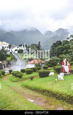 Furnas, Azores, Portugal - Jan 13, 2020: Volcanic hot springs in Portuguese Furnas. Steam coming from the water pools. Houses and hills in the background. Christmas decoration, green bushes and trees. Stock Photo