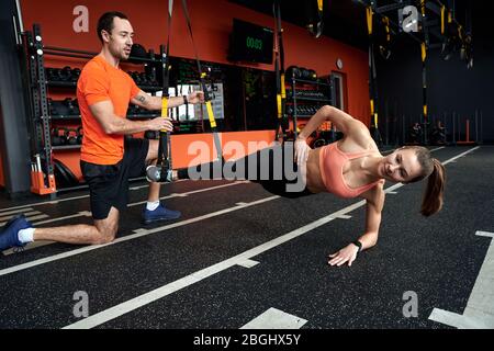 Sporty Lifestyle. Coach training and motivating young smiling woman doing side-plank tap TRX exercise at gym haging Stock Photo
