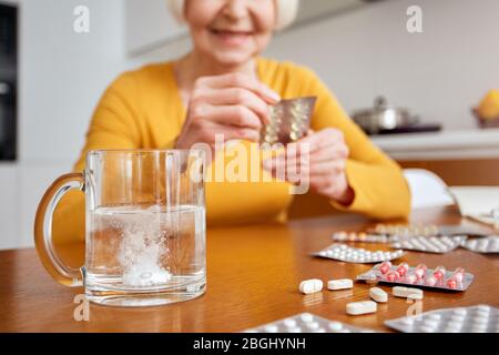 Retirement. Senior woman sitting at table at kitchen dissolving tablet in water close-up smiling cheerful blurred Stock Photo